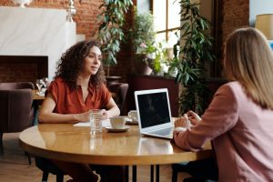 woman sat at public coffee table opposite woman with laptop