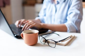 Remote working from home. Freelancer workplace in kitchen with laptop, cup of coffee, spectacles. Concept of distance learning, isolation, female business, shopping online. Close up of woman hands.