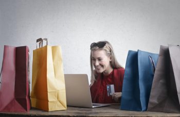 woman sits at table looking at laptop surrounded by bags of shopping