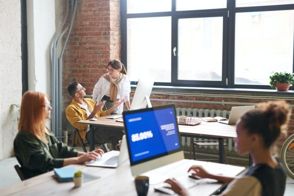 Office workers talk whilst sat at tables in front of large modern computers