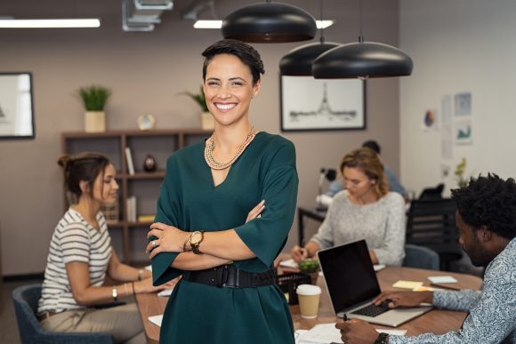 woman stood up smiling to camera while office workers sit around at a table working behind her