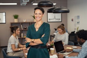 Portrait of successful business woman standing with her colleagues working in background at office. Portrait of cheerful fashion girl in green dress standing with folded arms and looking at camera. Beautiful businesswoman feeling proud and smiling.